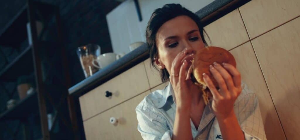 common signs of bulimia nervosa - woman eating burger on kitchen floor